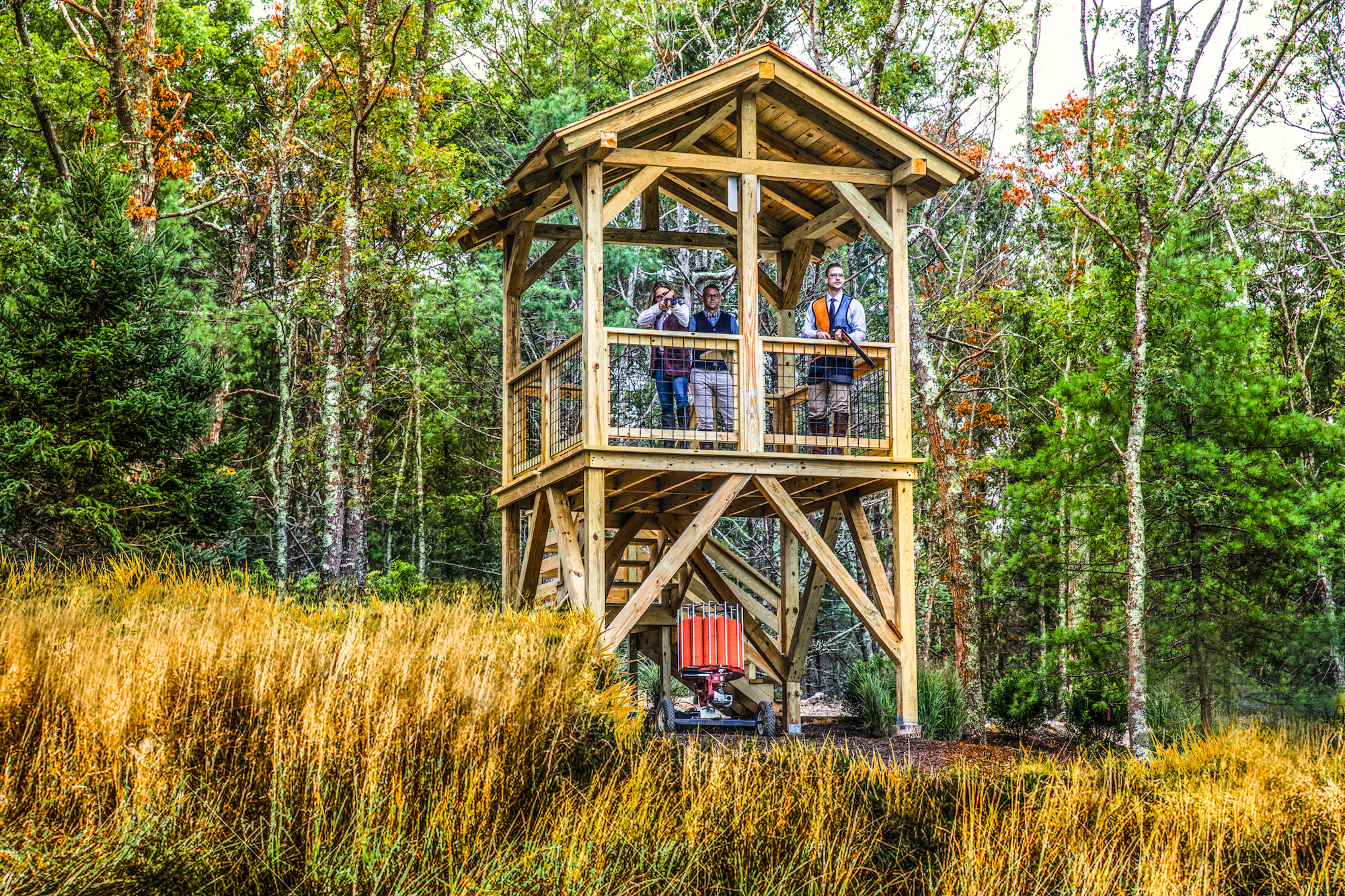 A wooden observation tower in a forested area, with several people on the upper platform looking out at the surroundings amidst tall grass and trees.