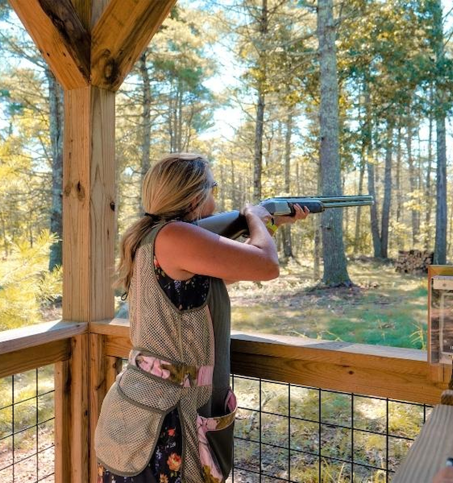 A person is aiming a shotgun in an outdoor setting while standing under a wooden structure, with trees visible in the background.