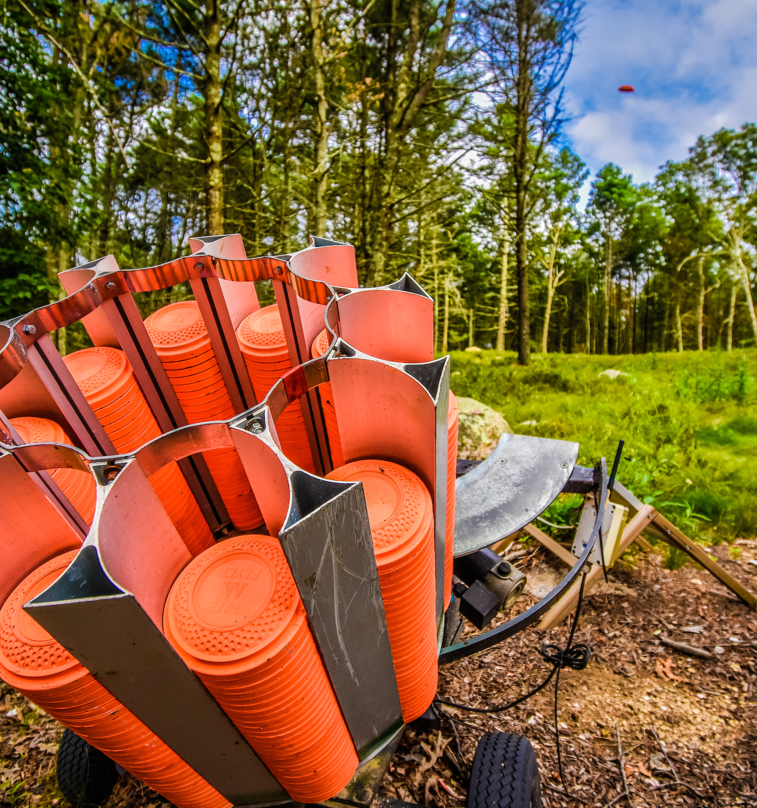 The image shows a clay pigeon shooting machine loaded with orange clay targets, set in a forested outdoor setting.