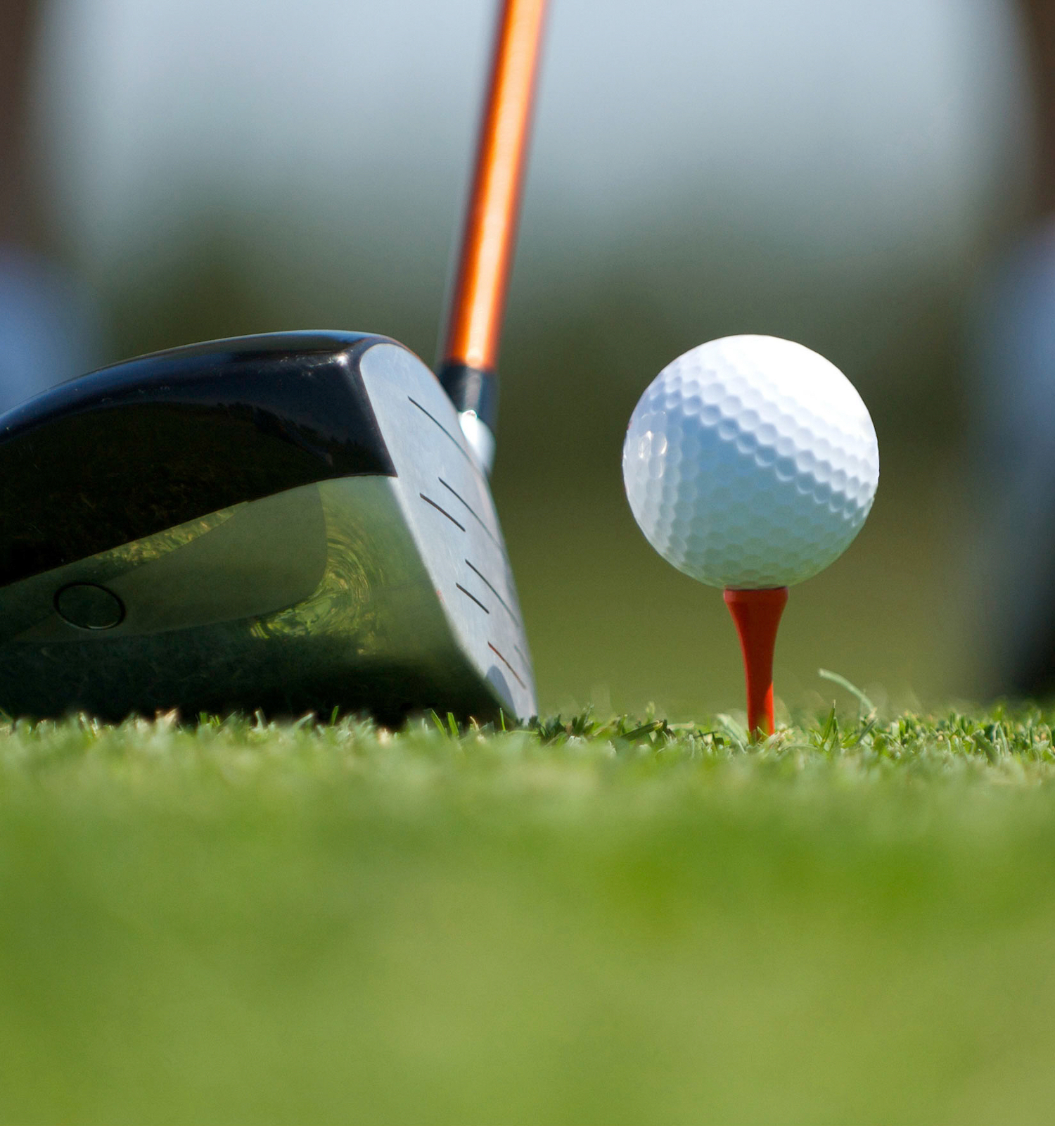 The image displays a golf club positioned to hit a golf ball that is teed up on a red tee on a golf course.