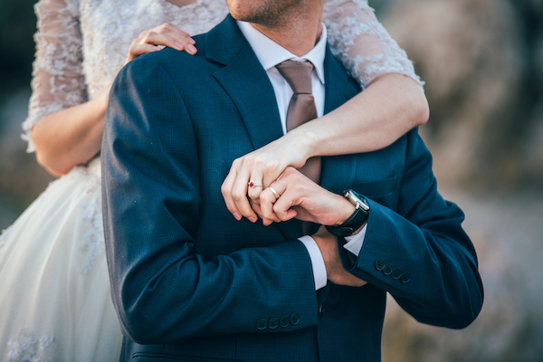 A groom in a blue suit is embraced by a bride in a white dress, showing their wedding rings.