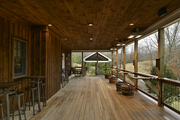 An outdoor wooden deck with bar stools, tables, and overhead lighting. It overlooks a scenic area with trees and a small wooden structure.