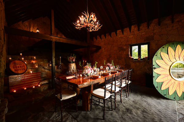 A cozy dining room with a rustic chandelier, a long wooden table set for a meal, and a vibrant sunflower mural on the right side.