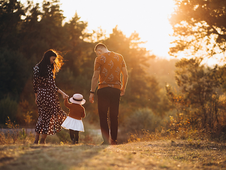 A family of three, with a young child, walking in a scenic, sunlit forest or field during sunset, holding hands and enjoying a peaceful moment together.