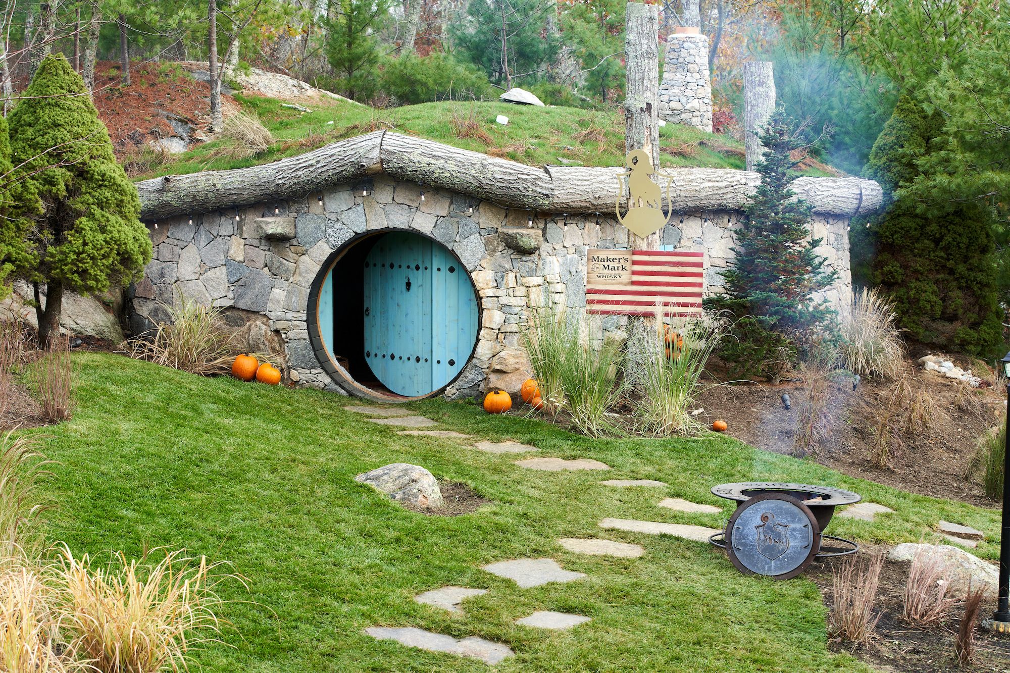 This image shows a Hobbit-style house with a round blue door, a stone path, pumpkins, and an American flag on a grassy slope, surrounded by trees.