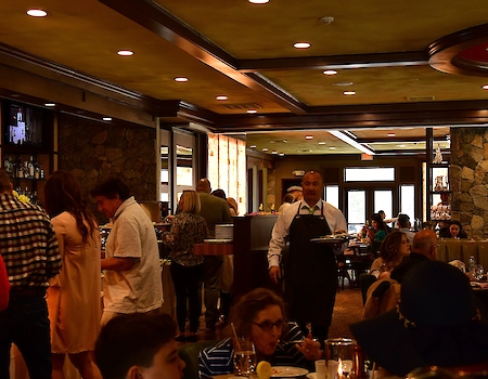 People are dining in a busy, warmly-lit restaurant with a waiter serving food and drinks to the tables, and a horse statue is on display.