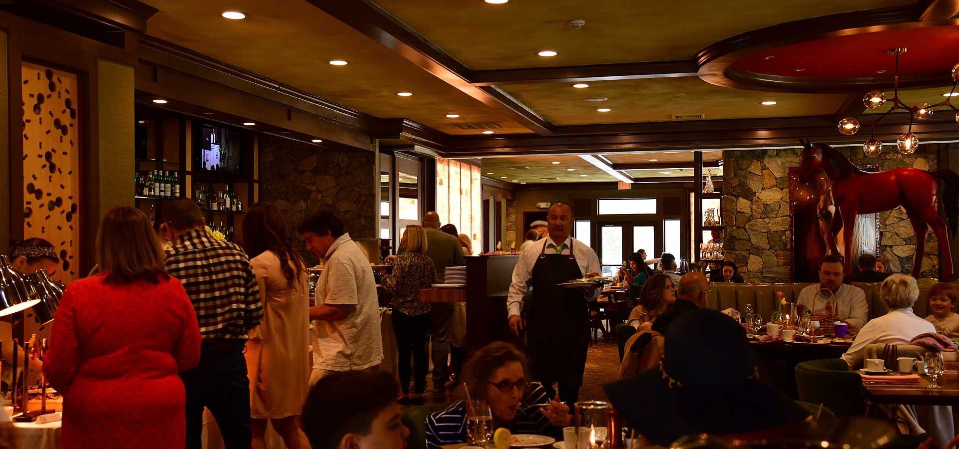 People are dining in a busy, warmly-lit restaurant with a waiter serving food and drinks to the tables, and a horse statue is on display.