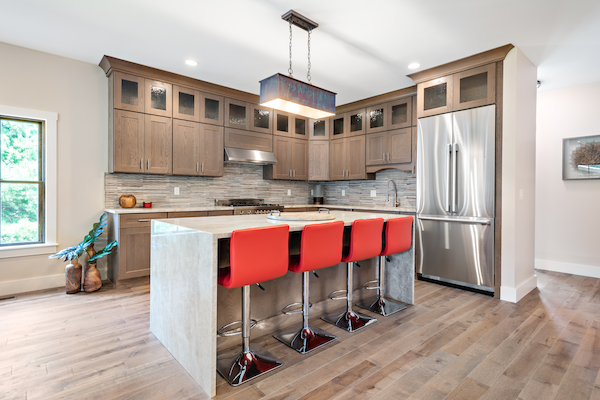 A modern kitchen with wooden cabinets, stainless steel appliances, an island with four red barstools, and wooden flooring is featured in the image.