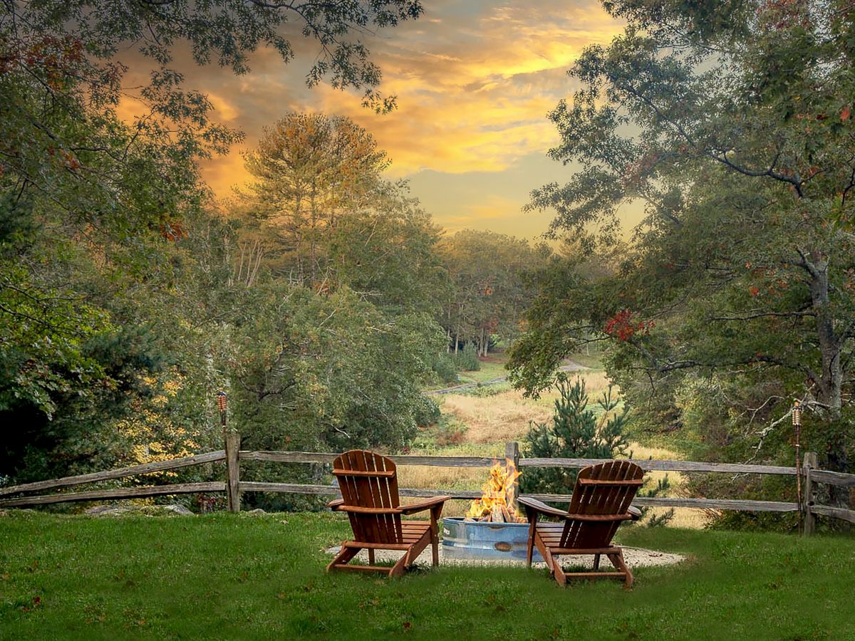 Two wooden chairs sit by a fire pit in a lush, green backyard with a wooden fence, overlooking a wooded area at sunset.