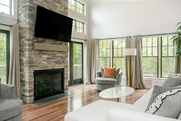 A modern living room features large windows, a stone fireplace, TV, gray furniture, a white coffee table, and cozy decor elements.