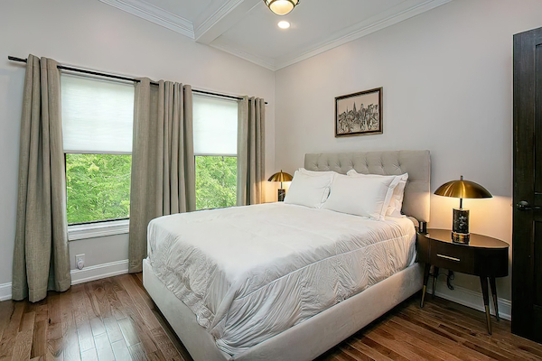 A cozy bedroom with a neatly made bed, two bedside tables with lamps, curtains, and hardwood floors. Wall art hangs above the bed.