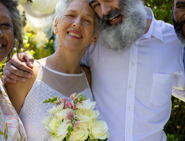 A joyful couple poses together, with the woman holding a bouquet of flowers, joined by two other people in a sunny outdoor setting.