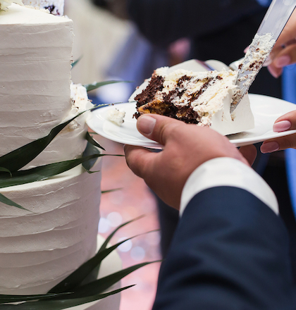 A person is serving a slice of cake from a tiered, white frosted cake decorated with green leaves onto a plate.