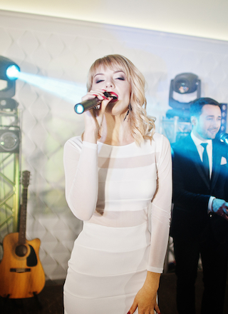 A woman in a white dress is singing into a microphone on stage. A man in a suit is in the background with a guitar visible nearby.