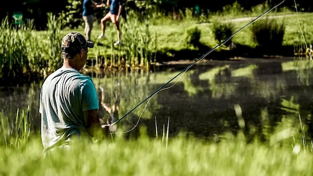 A person is fishing by a pond, surrounded by grass, with two more people walking in the background.