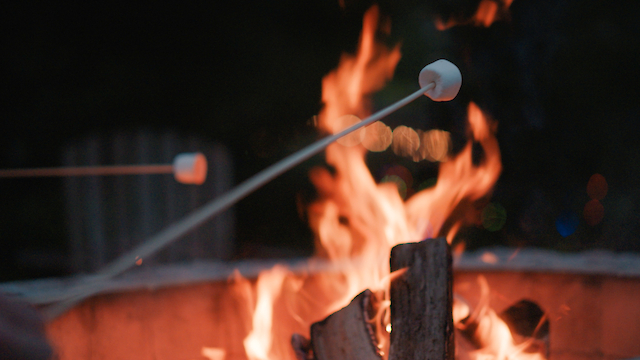 Two marshmallows on sticks being roasted over an open outdoor fire pit, against a dark background.