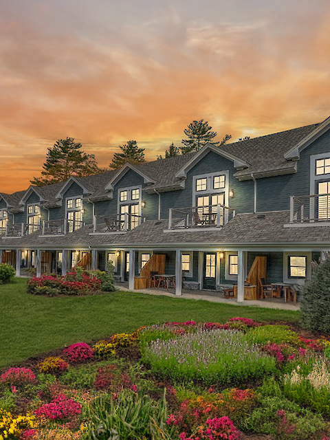 The image shows a row of charming cottages with balconies, surrounded by lush gardens and a vibrant sunset sky.