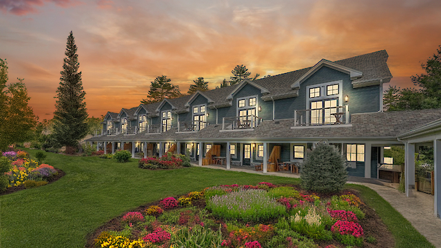 The image shows a row of charming cottages with balconies, surrounded by lush gardens and a vibrant sunset sky.