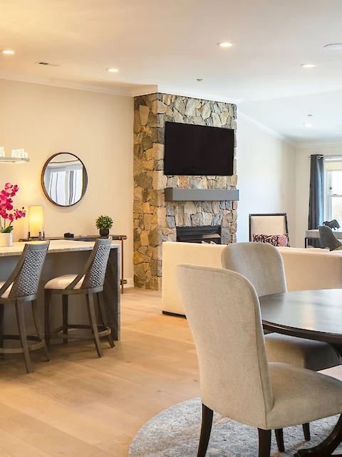 A modern open-plan living space with a kitchen, dining area, and cozy seating. Features neutral tones, warm lighting, a TV, and wall art.