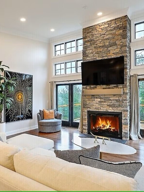 A modern living room with a fireplace, TV, large windows, and comfortable seating, featuring a decorative plant and artwork on the wall.