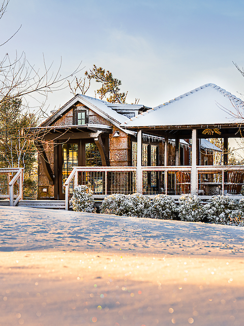 A cozy cabin with a wooden deck is covered in snow, surrounded by bare trees and a snowy landscape, under a clear sky.