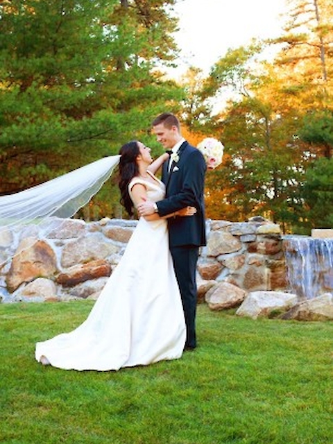 A couple in wedding attire poses outdoors on grass, near rocks and trees, with a long veil flowing behind the bride.