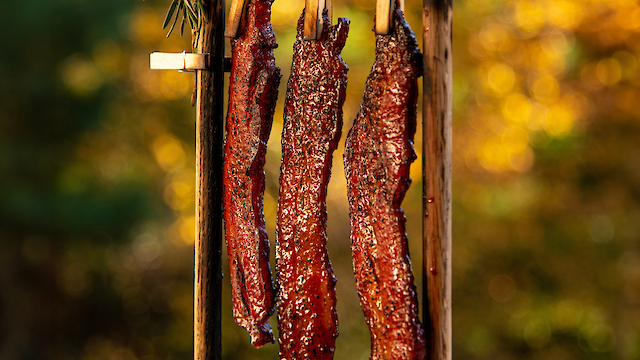 Three strips of cooked bacon hang from clothespins on a wooden frame, with a pickle and lemon wedge at the base.