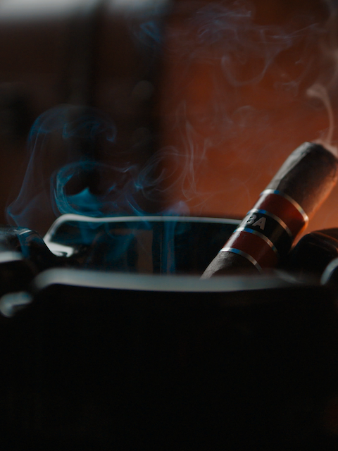 A lit cigar rests in an ashtray with smoke rising, surrounded by glasses of whiskey, creating a relaxing atmosphere.