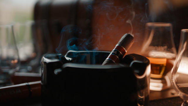 A lit cigar rests in an ashtray with smoke rising, surrounded by glasses of whiskey, creating a relaxing atmosphere.