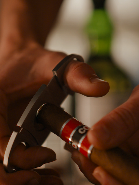 A person is using a cigar cutter to clip the end of a cigar. There’s a bottle in the blurred background.