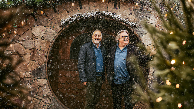 Two people stand in front of a round doorway framed by a stone wall, amidst falling snow and surrounded by trees with lights.