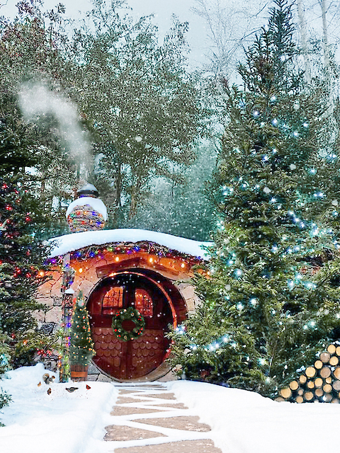 A cozy, snow-covered house in a forest with decorative lights and smoke from the chimney, surrounded by snowy trees and a woodpile.