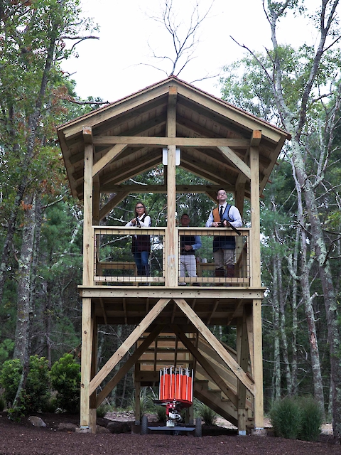 A wooden observation tower in a forest with two people standing on the top level. There's an object with red sections beneath the tower.