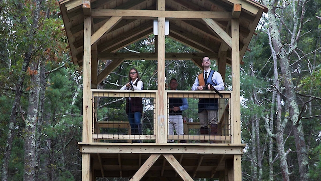 A wooden observation tower in a forest with two people standing on the top level. There's an object with red sections beneath the tower.