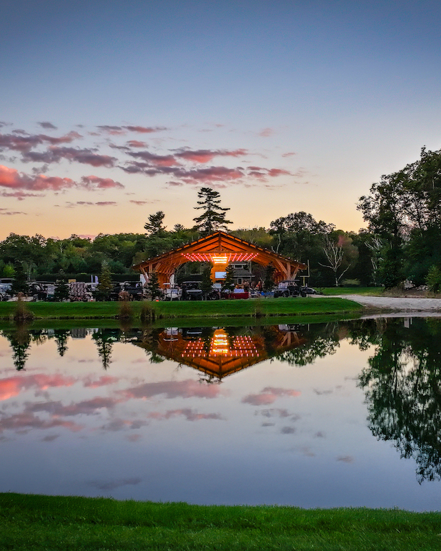 A picturesque lakeside scene at sunset with a wooden pavilion lit warmly, reflecting in the water, surrounded by trees and a vibrant sky.